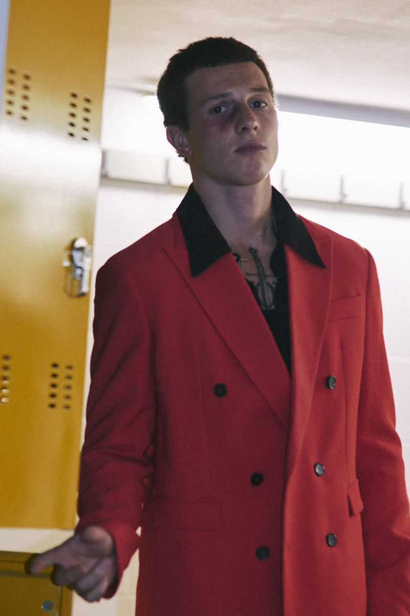 Young man wearing a bright red double-breasted blazer and black open-collar shirt standing beside a yellow locker.