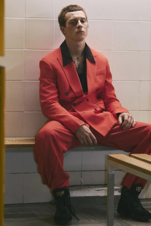Young man wearing a bright orange suit and black sneakers seated against a white tiled wall for CRUSHfanzine.