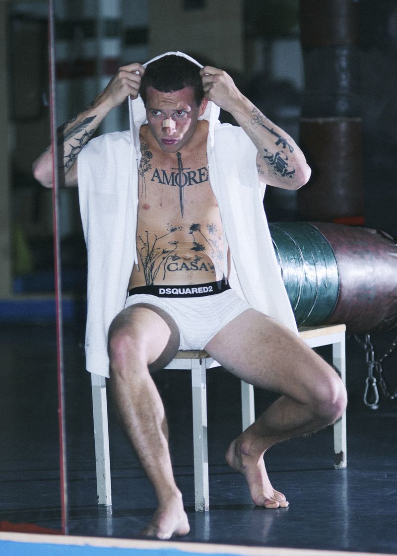 Tattooed man in white briefs and a white sleeveless hoodie seated on a chair in a boxing gym with punching bags in the background.