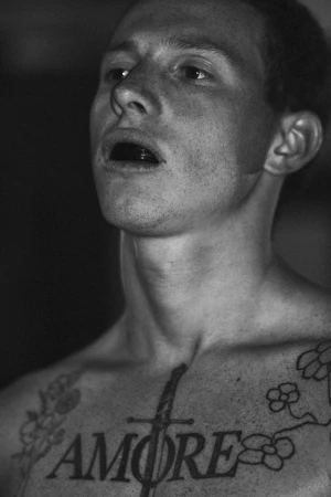 Black and white close up portrait of a male fighter with chest tattoos and a black mouthguard, looking upward against a dark background.