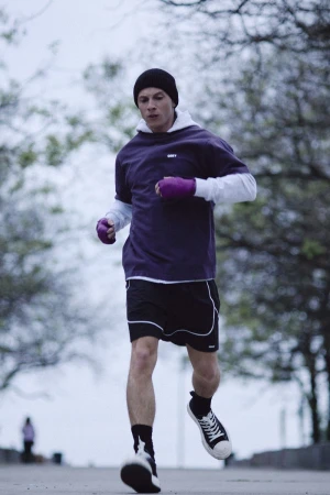 A man running outdoors wearing a purple t-shirt, black beanie, purple hand wraps, and athletic shorts along a tree-lined path.