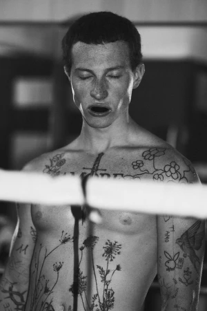 Black and white portrait of a shirtless man with extensive chest and arm tattoos standing behind a boxing ring rope with eyes closed.