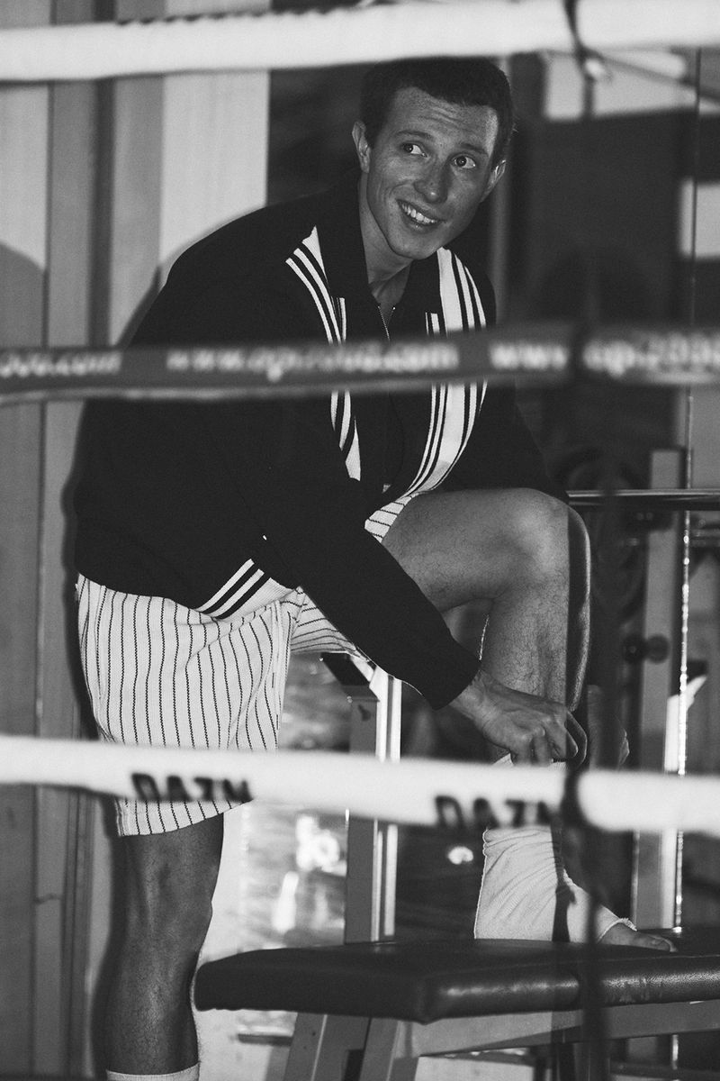 A smiling man adjusting ankle wraps on a bench inside a boxing ring, wearing a dark track jacket and striped shorts.
