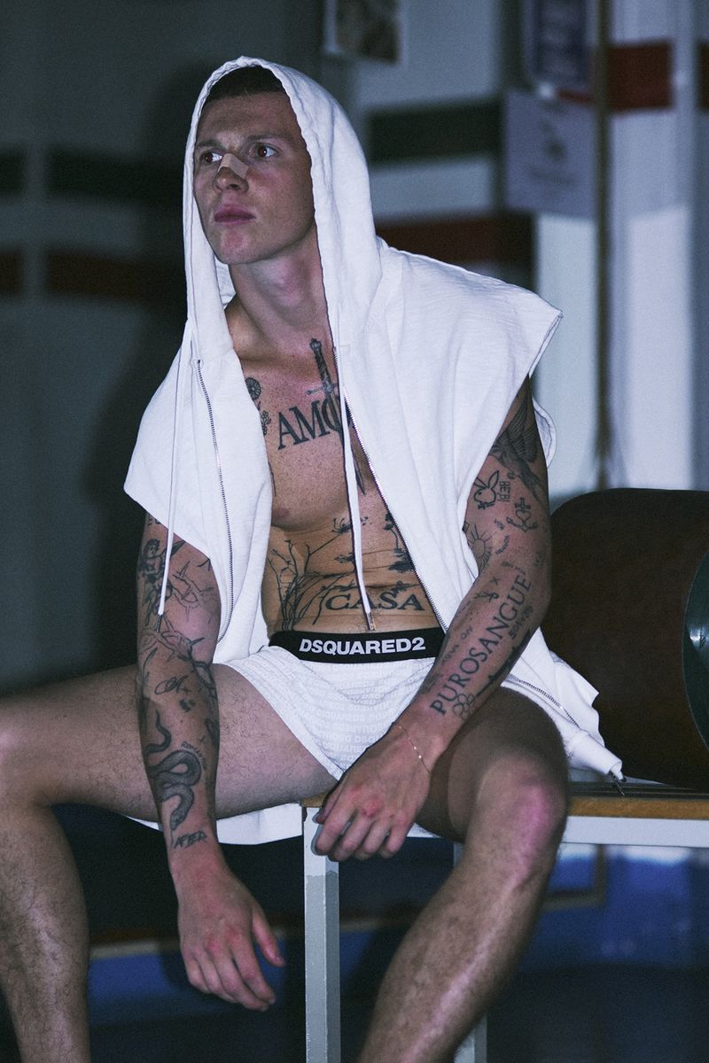 Tattooed man in a white hooded vest and boxer briefs with a nose bandage sitting on a bench indoors.