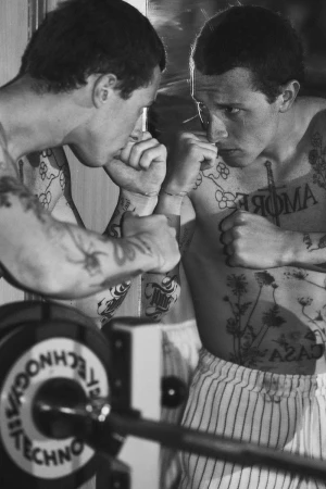 Black and white photo of a shirtless tattooed man raising his fists in front of a mirror in a gym.
