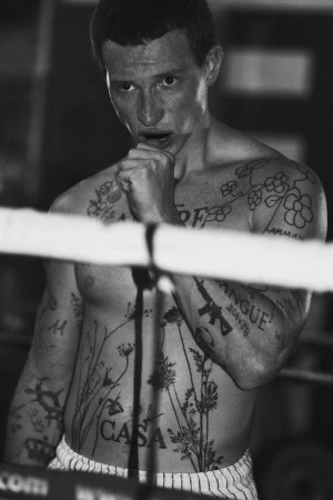 Black and white photograph of a tattooed man holding his fist to his chin inside a boxing ring.