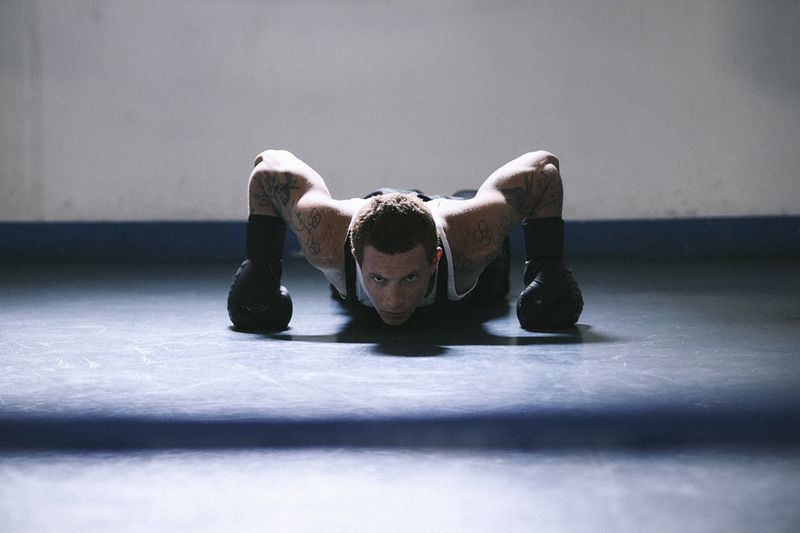 Man with arm tattoos wearing black boxing gloves and a white tank top in a low push-up position on a training floor.