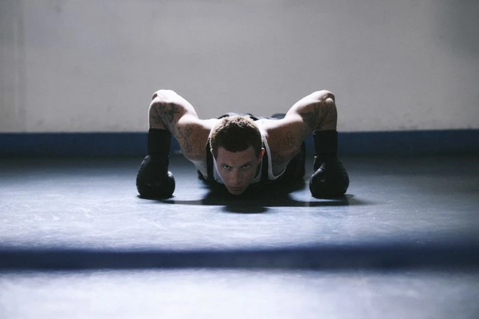 Man with arm tattoos wearing black boxing gloves and a white tank top in a low push-up position on a training floor.
