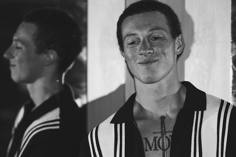 Black and white portrait of a smiling young man in a striped knit polo shirt with his reflection visible in a mirror.