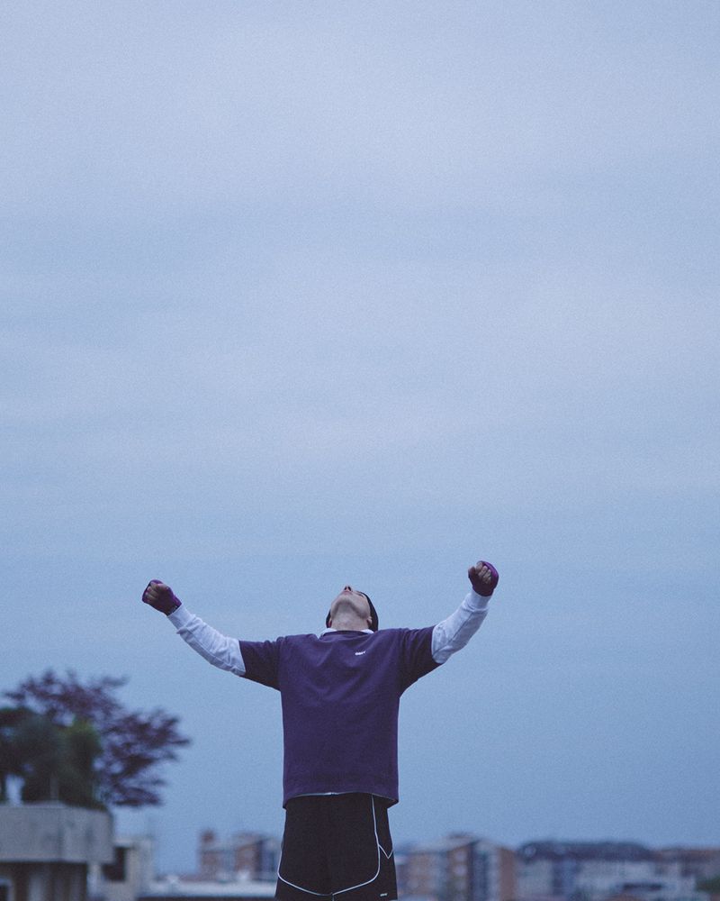 A man with taped hands stands outdoors looking up toward an overcast sky with both arms raised in celebration or victory.