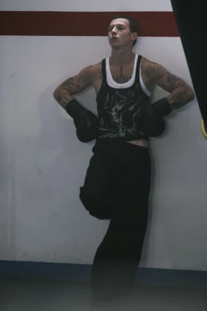 Tattooed man wearing boxing gloves, a graphic tank top, and black wide-leg pants leaning against a white wall with red trim.