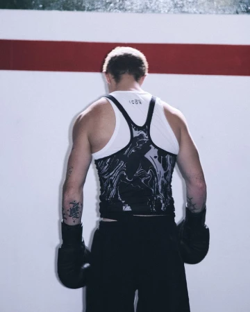Rear view of a boxer wearing black gloves, layered racerback tank tops, and shorts against a white wall with a red stripe.