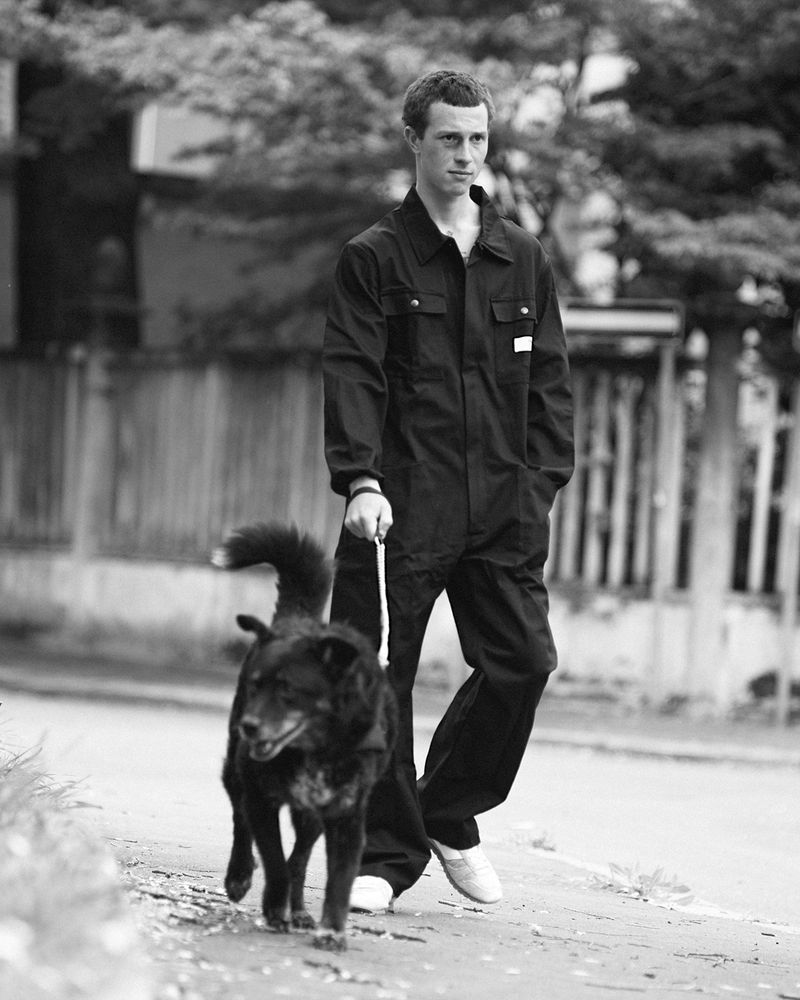 Man in a dark utility jumpsuit walking a black dog on a leash along a paved street in black and white.
