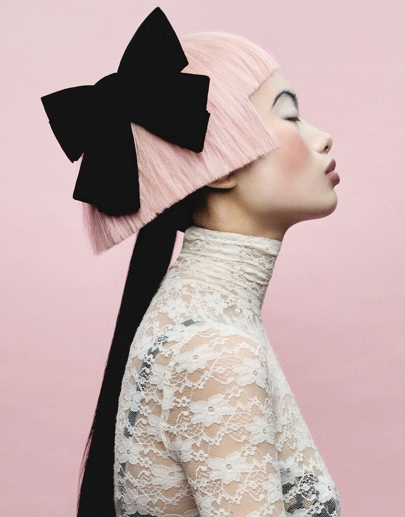 Model in profile with blunt pink hair, a large black hair bow, and a white lace turtleneck against a pink studio backdrop.
