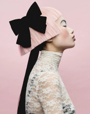 Model in profile with blunt pink hair, a large black hair bow, and a white lace turtleneck against a pink studio backdrop.