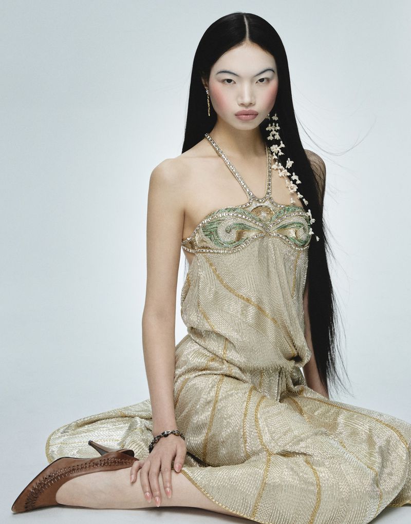 Model Chun Jin sits in a gold halter dress with long black hair adorned with floral clips against a white studio background.