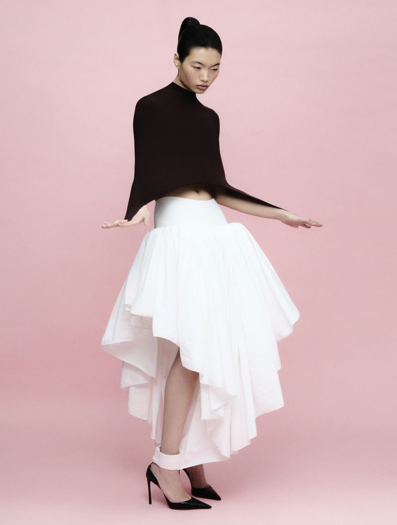 Chun Jin posing in a black structured cape top and voluminous white high-low skirt against a pink backdrop.