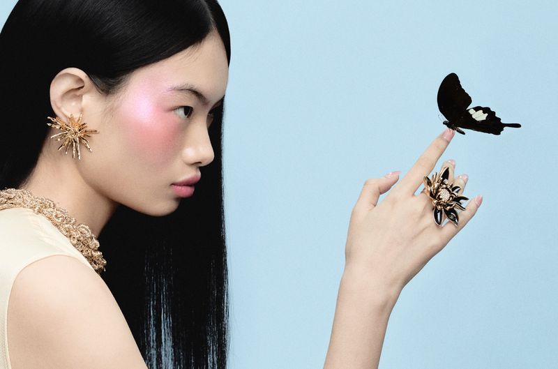 Model Chun Jin in profile with a black butterfly perched on her finger, wearing gold jewelry against a pale blue background.