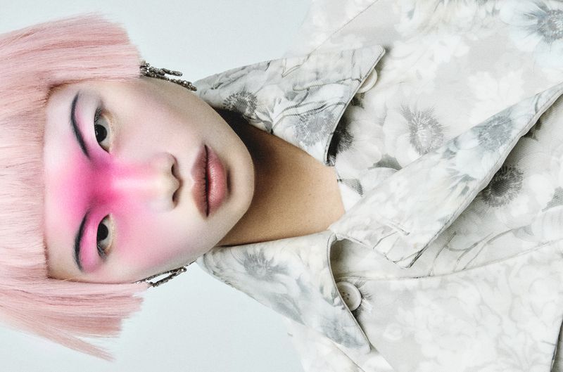 Model Chun Jin with a pastel pink bob and vivid pink eye makeup wearing a floral patterned trench coat against a white backdrop.