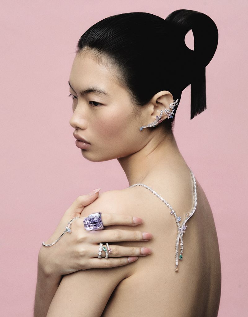 Model Chun Jin wearing high jewelry ear cuffs, rings, and necklace draped over bare shoulders against a pink backdrop.