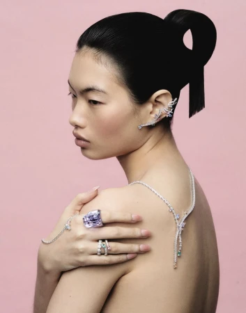 Model Chun Jin wearing high jewelry ear cuffs, rings, and necklace draped over bare shoulders against a pink backdrop.