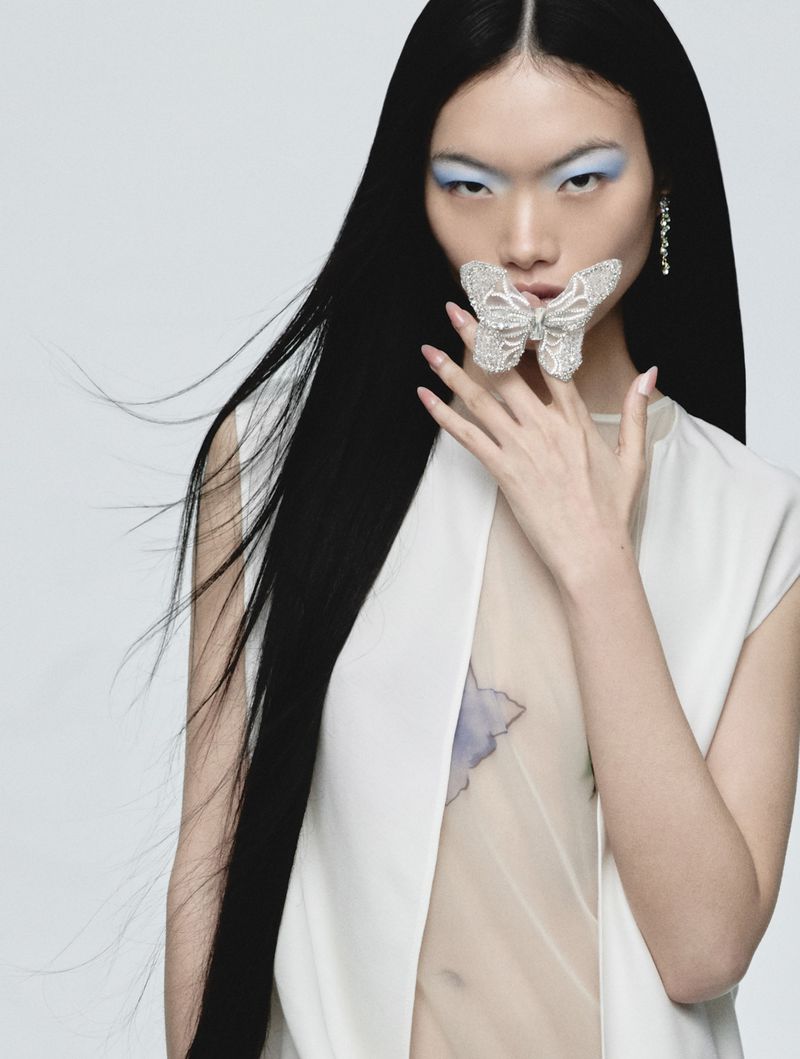 Model Chun Jin holds a shimmering silver butterfly accessory across her mouth in a studio portrait against a light backdrop.