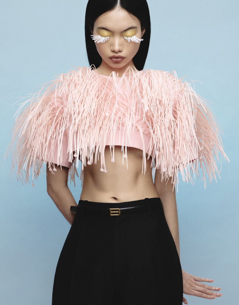 Model with long dark hair wearing a pink fringe top and black trousers against a light blue backdrop.