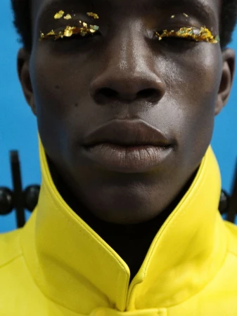 Close-up portrait of model Khalil Bayo with gold leaf eyelids wearing a bright yellow high-collar jacket against a blue backdrop.