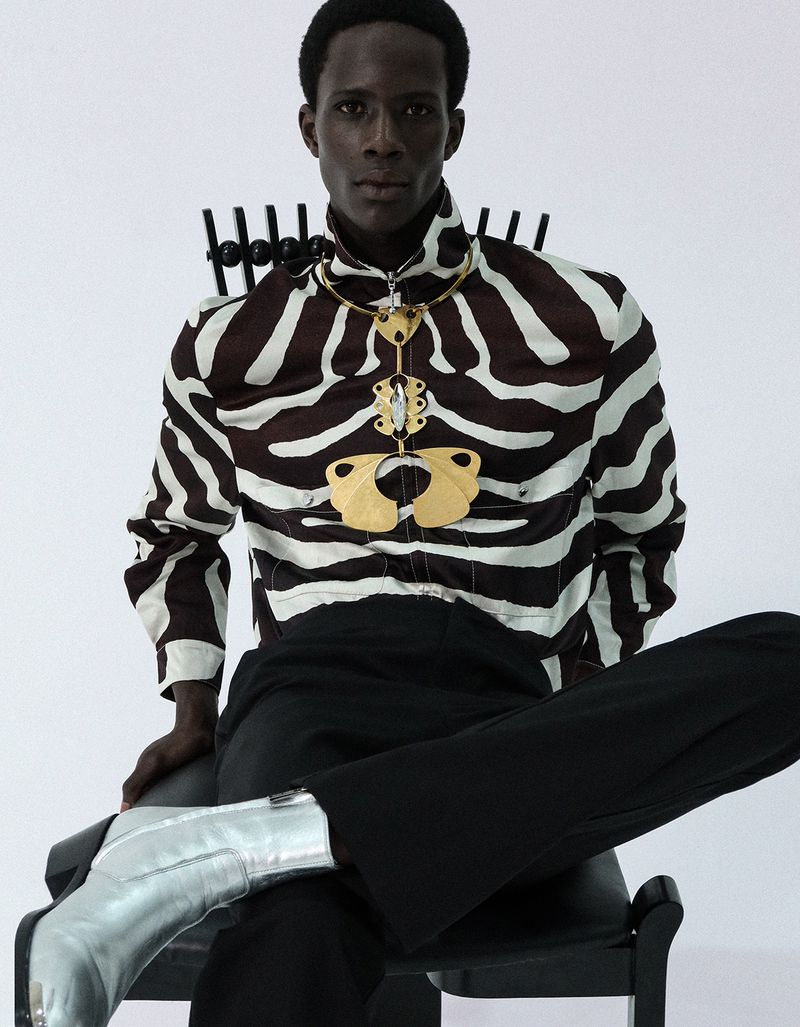 Black man seated on a chair wearing a zebra print zip shirt, large gold necklace, black trousers, and silver boots on white background.