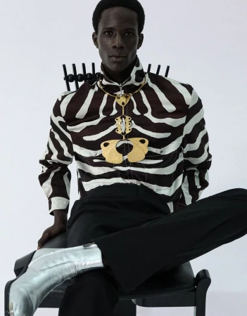 Black man seated on a chair wearing a zebra print zip shirt, large gold necklace, black trousers, and silver boots on white background.