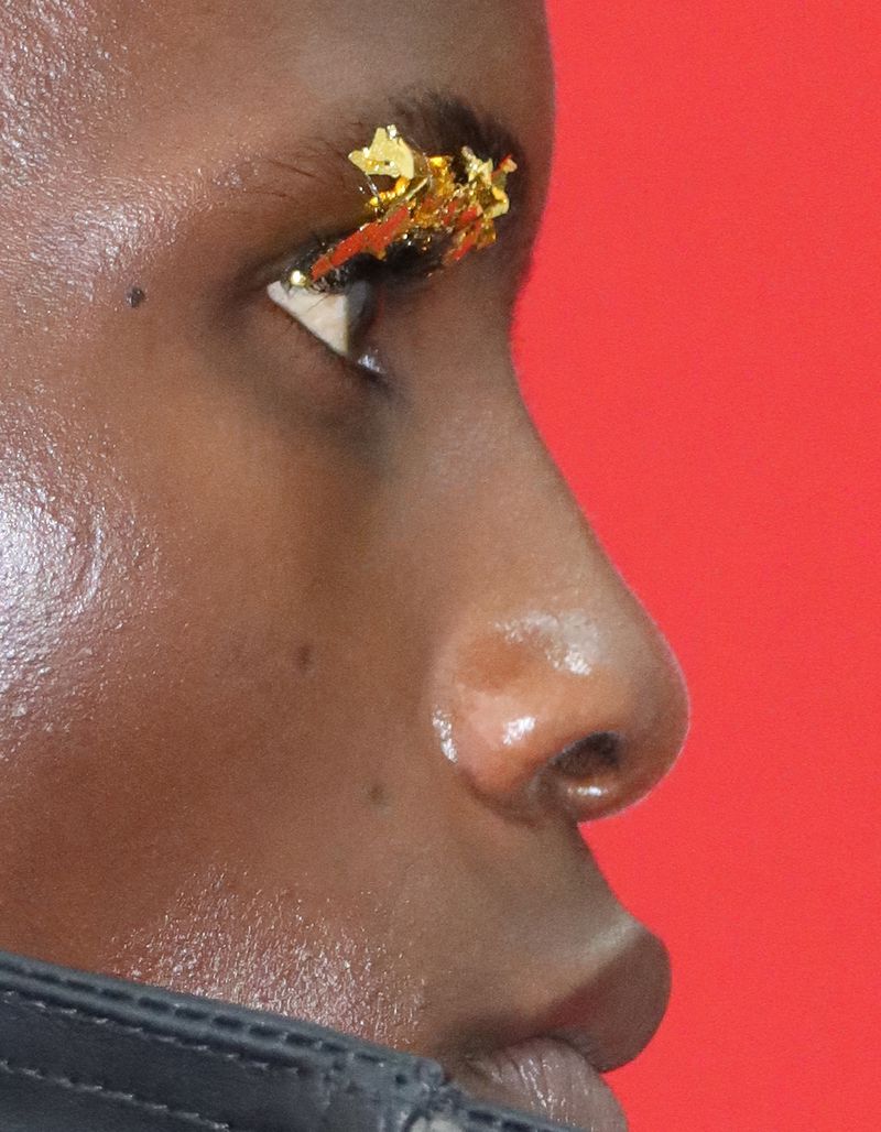 Profile close-up of a Black model with textured gold foil applied to the eyelid, set against a solid red studio background.