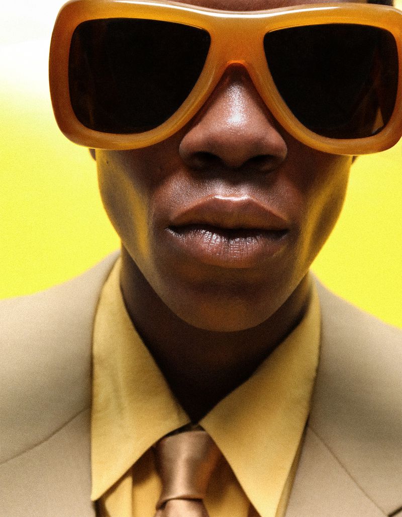 Model Khalil Bayo wearing chunky amber sunglasses, a beige suit jacket, yellow shirt, and brown tie against a yellow backdrop.