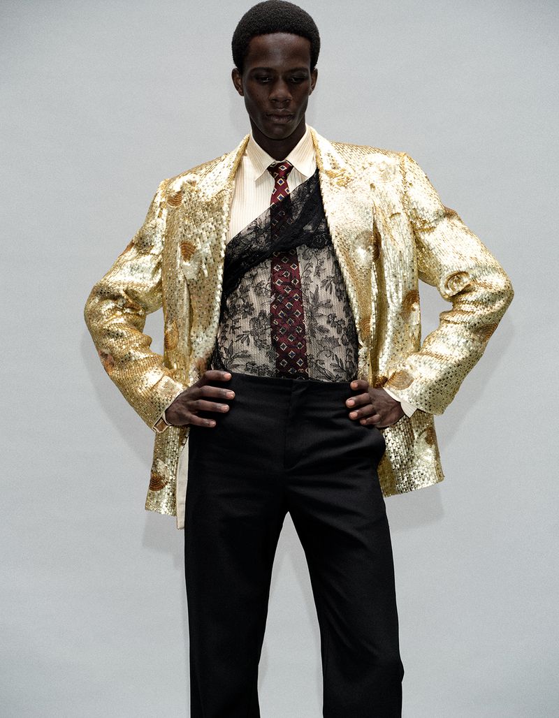 Model Khalil Bayo wearing a metallic gold sequin blazer, black lace overlay, and black trousers against a white studio backdrop.