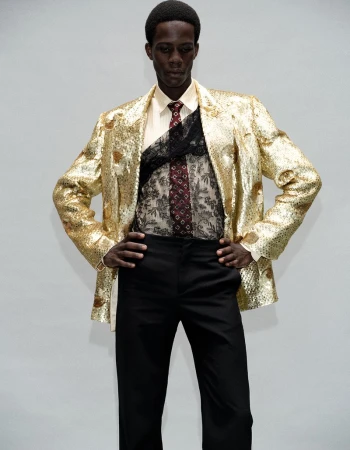 Model Khalil Bayo wearing a metallic gold sequin blazer, black lace overlay, and black trousers against a white studio backdrop.