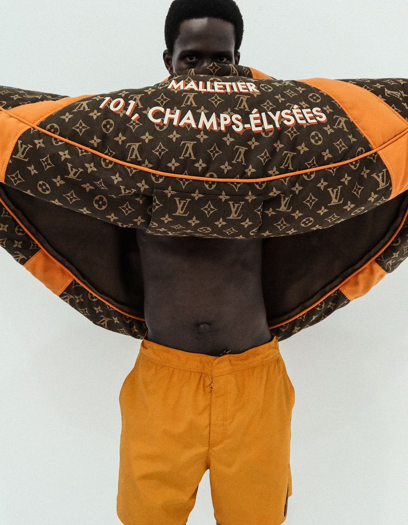 Model Khalil Bayo holds up a brown monogram and orange jacket across his face, wearing orange shorts against a white studio background.