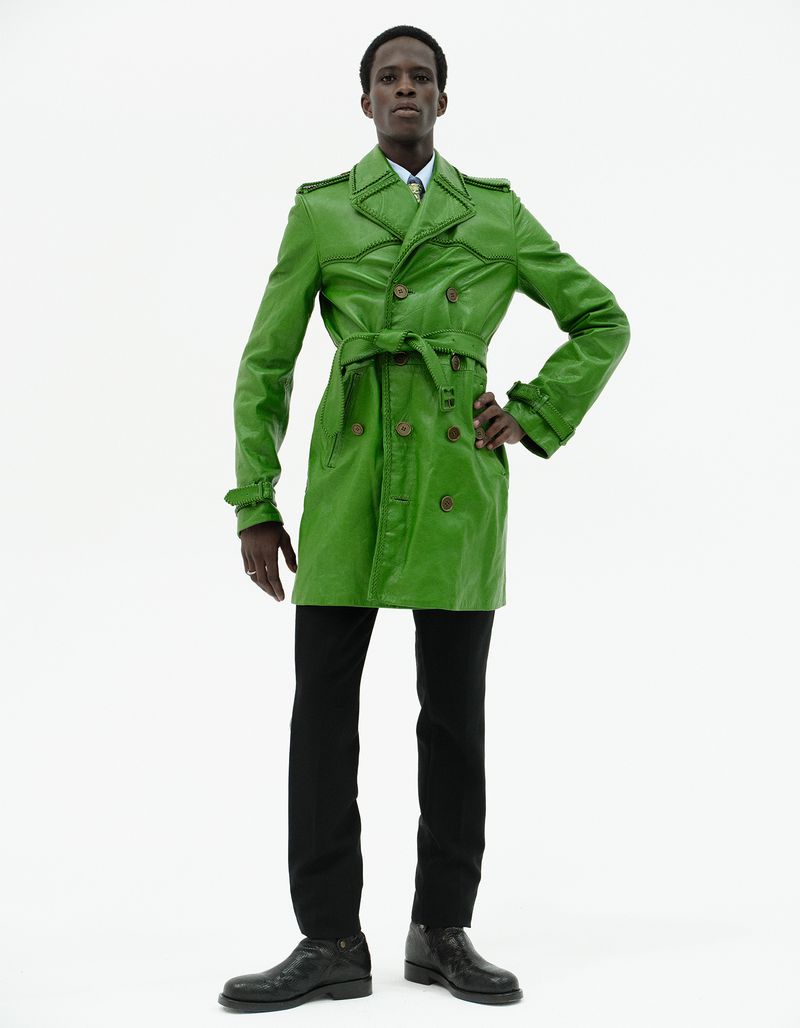 Model in a bright green leather trench coat and black trousers standing against a seamless white background.