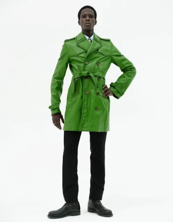 Model in a bright green leather trench coat and black trousers standing against a seamless white background.