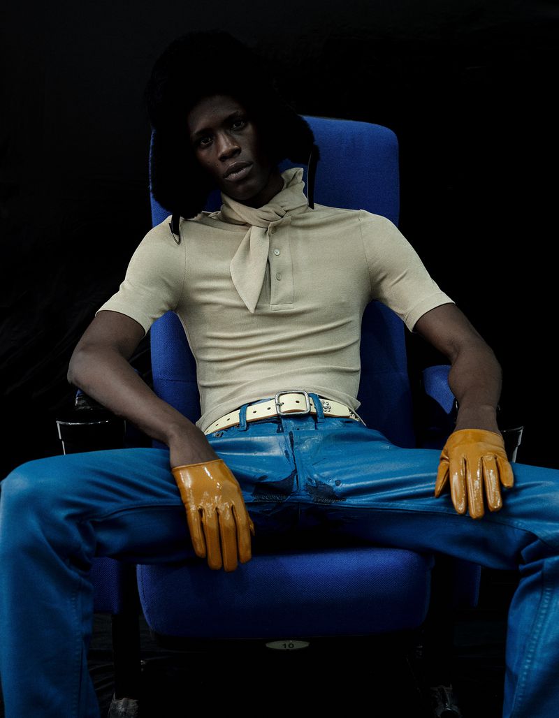 Model Khalil Bayo wearing a beige polo, blue pants, yellow gloves, and a black fur hat, seated in a blue chair against a black backdrop.