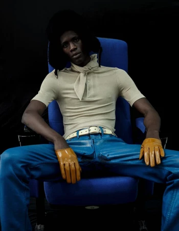 Model Khalil Bayo wearing a beige polo, blue pants, yellow gloves, and a black fur hat, seated in a blue chair against a black backdrop.