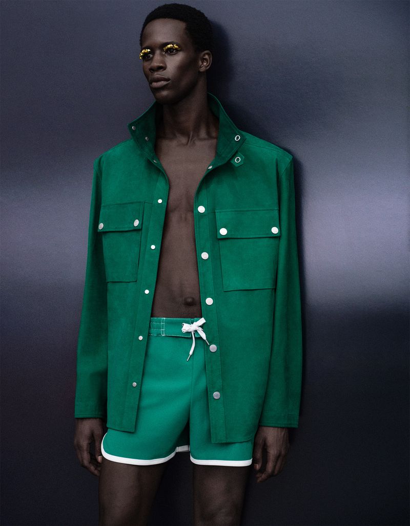 Model Khalil Bayo wearing an open green jacket and matching shorts with yellow eye makeup against a dark studio background.