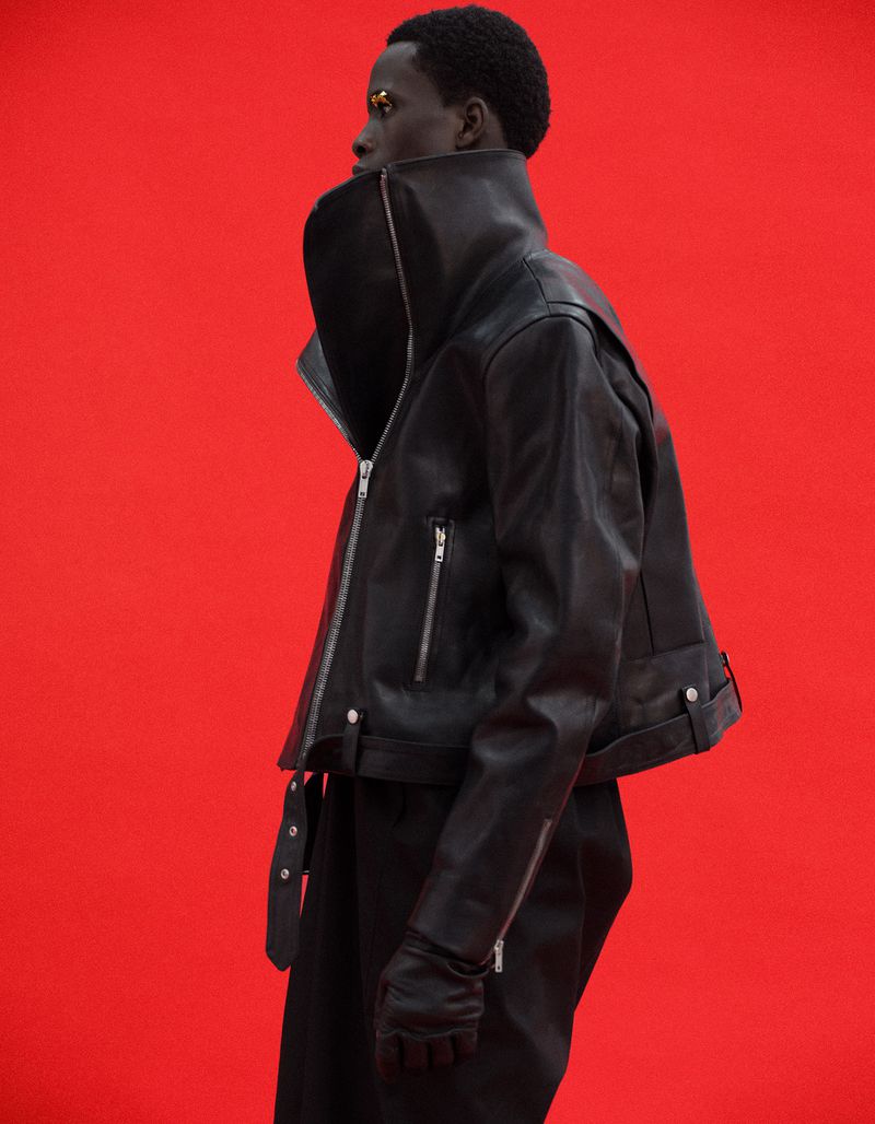 Model Khalil Bayo wearing a high-collar black leather cropped biker jacket with silver zippers against a bright red studio backdrop.