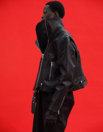 Model Khalil Bayo wearing a high-collar black leather cropped biker jacket with silver zippers against a bright red studio backdrop.