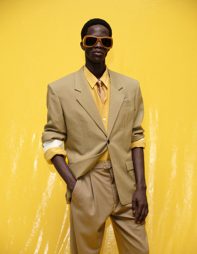 Model Khalil Bayo posing in an oversized beige suit, yellow collared shirt, and matching sunglasses against a bright yellow studio backdrop.