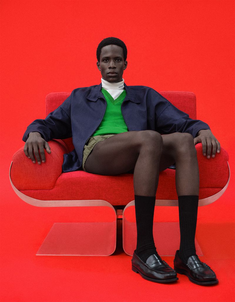 Model Khalil Bayo sits in a red armchair against a vivid red seamless background wearing a navy jacket and emerald green sweater.