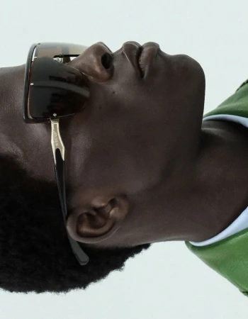 Profile view of a Black man wearing dark wrap-around sunglasses and a green collared jacket against a pale studio background.