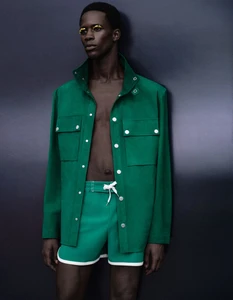 Model Khalil Bayo wearing an open green jacket and matching shorts with yellow eye makeup against a dark studio background.