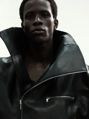 Model Khalil Bayo wearing an oversized black leather jacket with an exaggerated high collar against a white background.