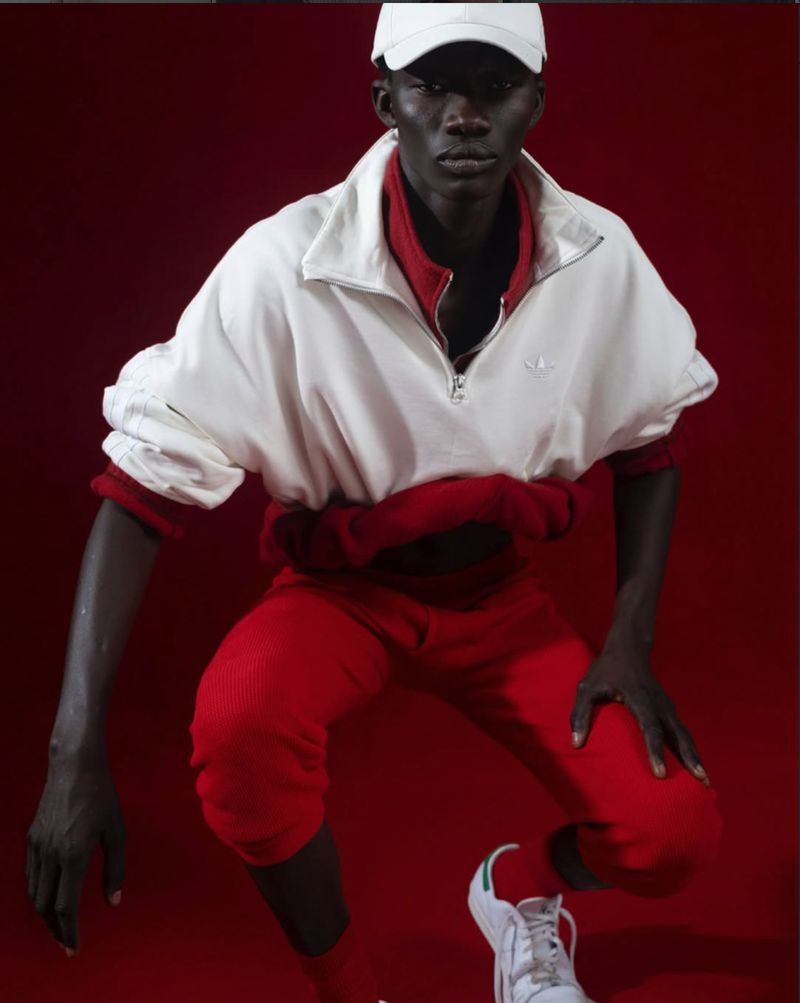 Model Saliou Gueye crouched in a white zip pullover, red pants, white cap, and white sneakers against a deep red background.