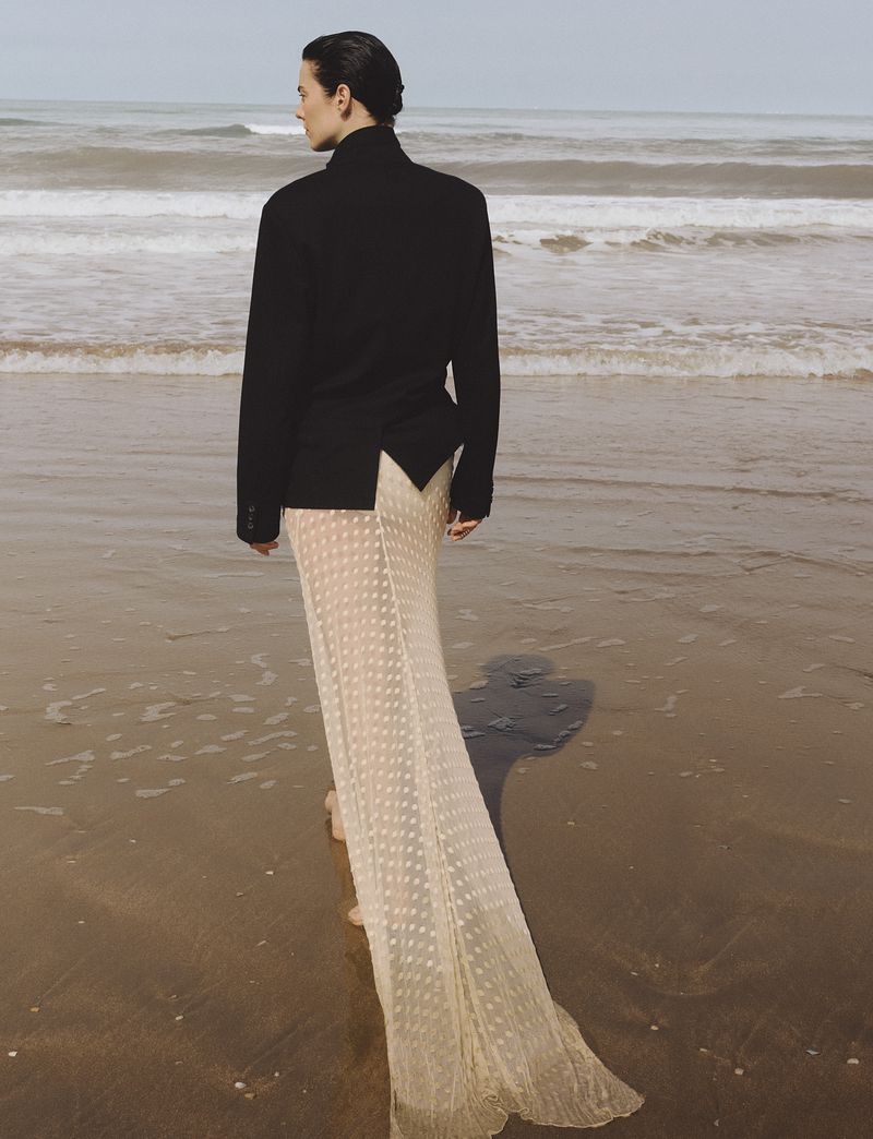 Model standing on a wet sandy beach facing the ocean wearing a black tailored blazer and sheer cream floor-length train skirt.