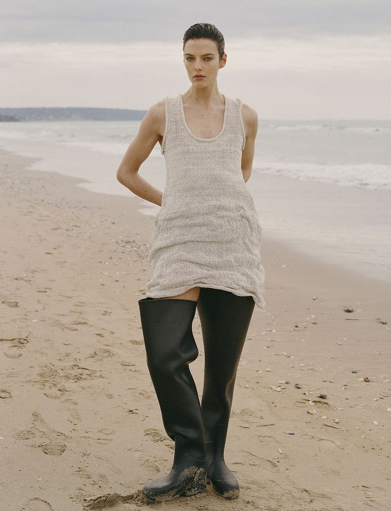 Model Beauïse Genç in a sleeveless knit tunic and black wader boots standing on a sandy beach for Numéro France.